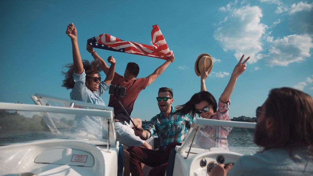 Group of friends on a boat with one holding an American flag, enjoying a sunny day.