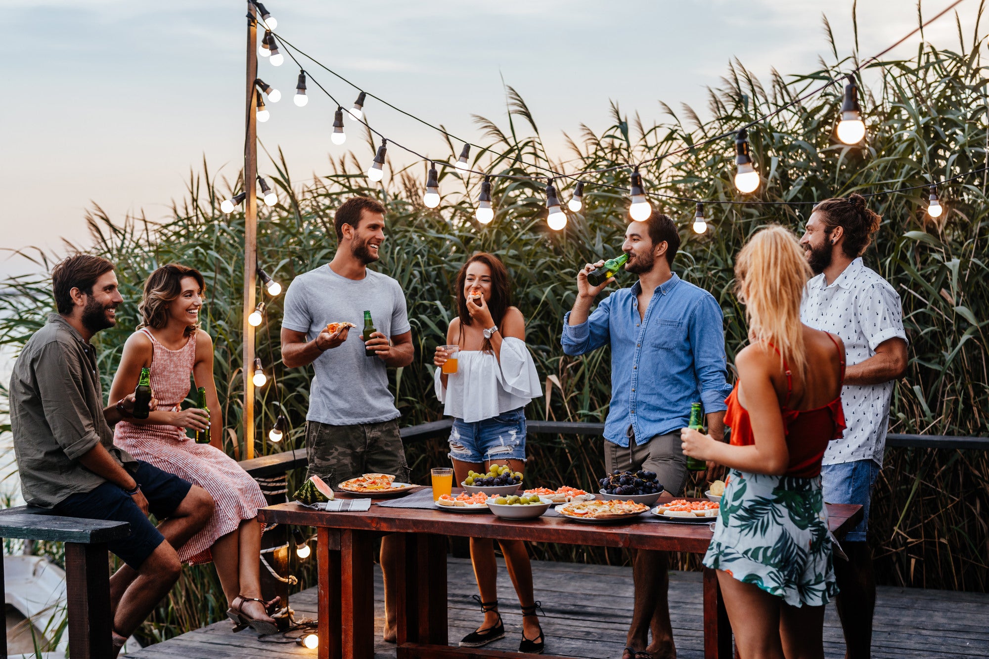 Group of friends socializing around a table with food and drinks on a rooftop with cornfield view.