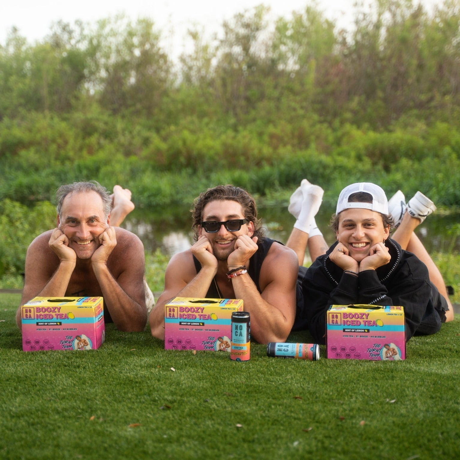 Three people lying on grass with boxes of Too Turnt Tea product in front of them.